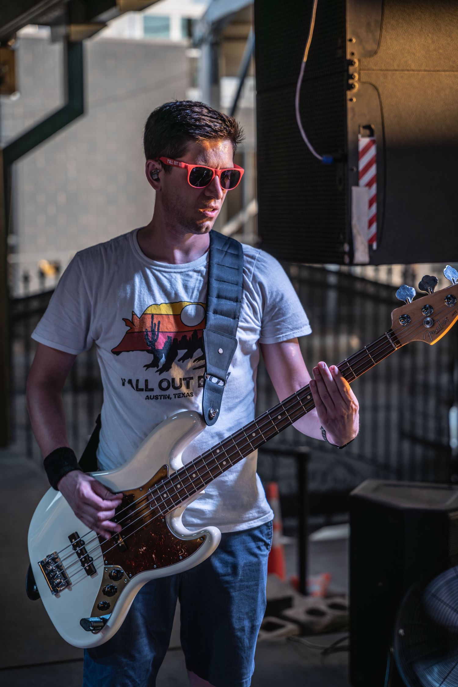 Musician wearing red sunglasses plays white bass guitar outdoors.