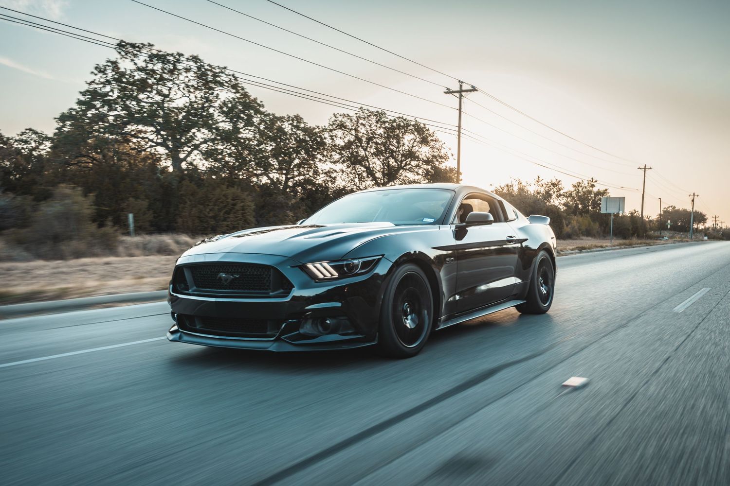 Dark Ford Mustang GT with black wheels cruising on a highway at dusk.