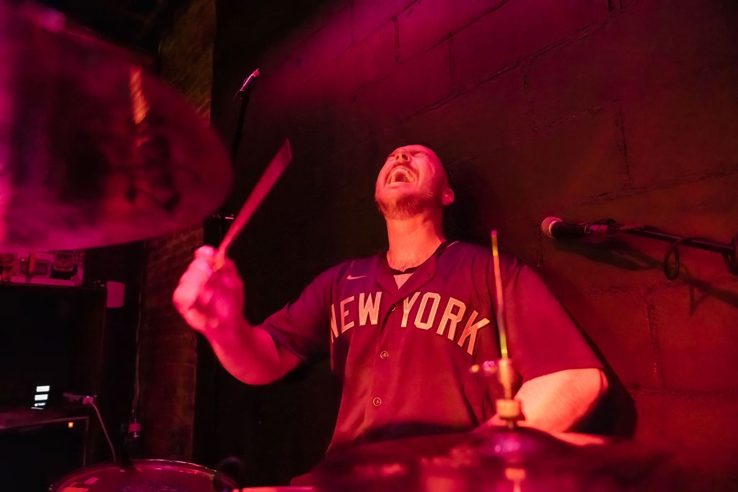 Drummer performing in New York shirt under dramatic red stage lighting.