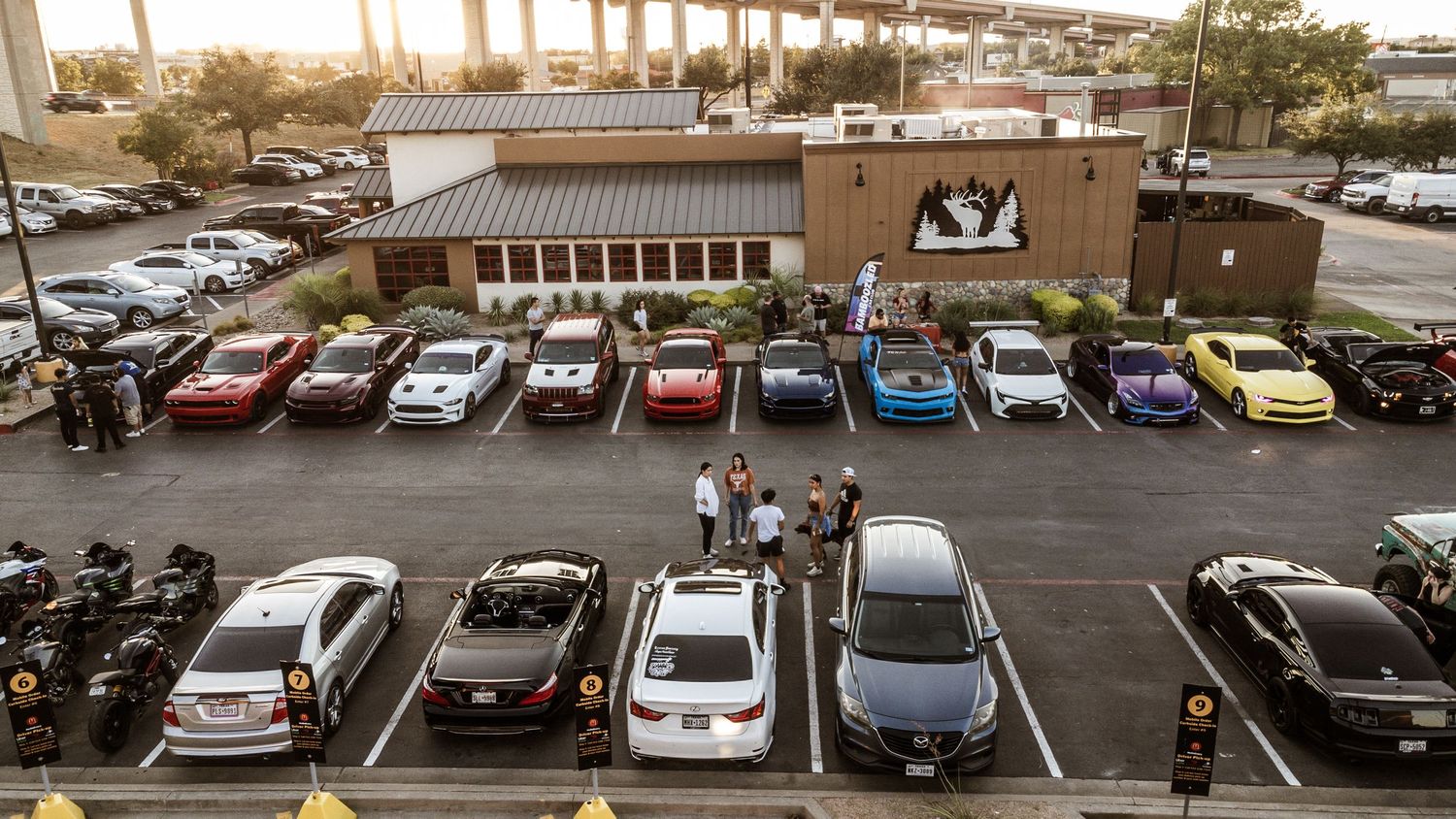 Car meet gathering in a parking lot with various modified vehicles lined up at dusk.