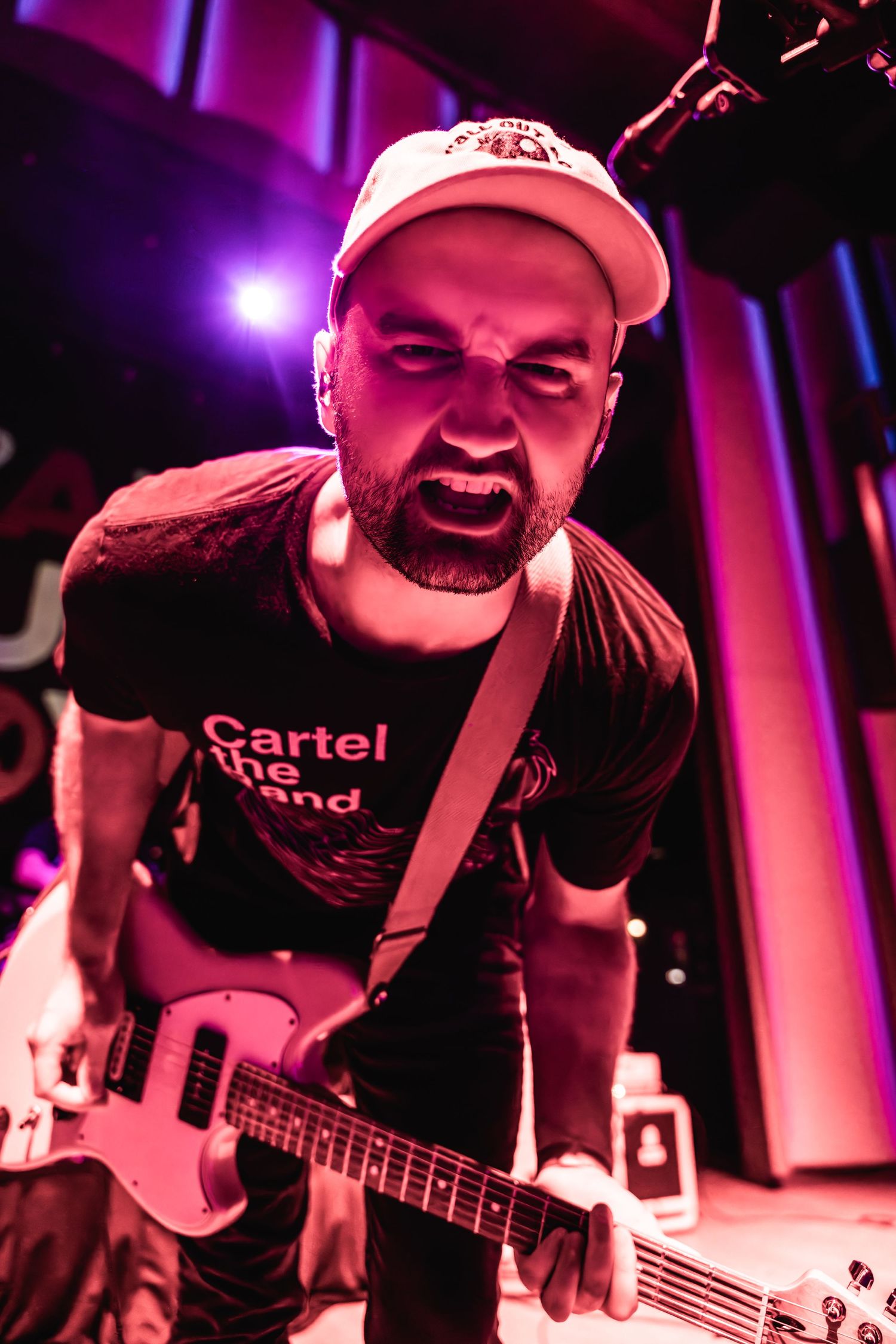 Bass player in cap and Cartel shirt performing passionately under red stage lights.