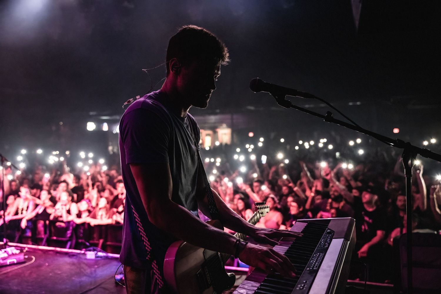 Silhouetted performer plays keyboard against backdrop of glowing phone lights from audience.