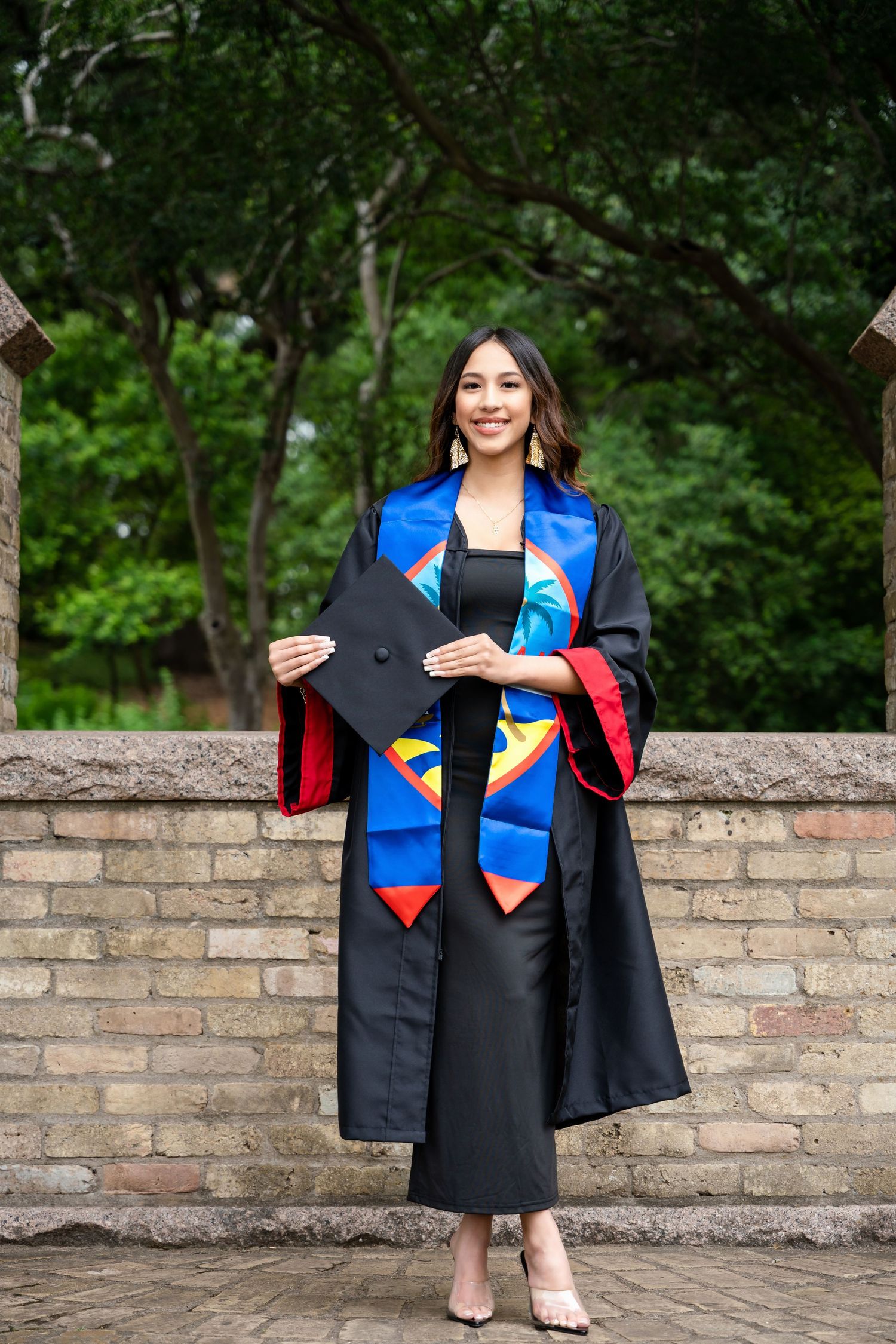 Graduate wearing black academic regalia with colorful stoles and sashes poses by brick wall in outdoor setting.