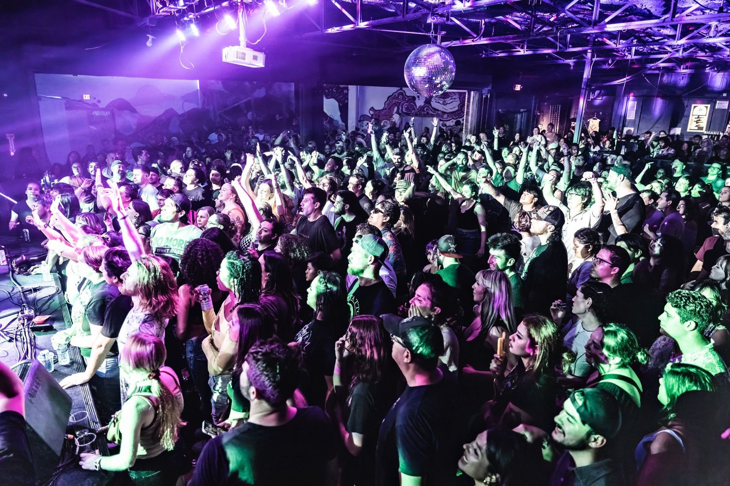 Large crowd fills a nightclub dance floor under colorful purple and green lighting.