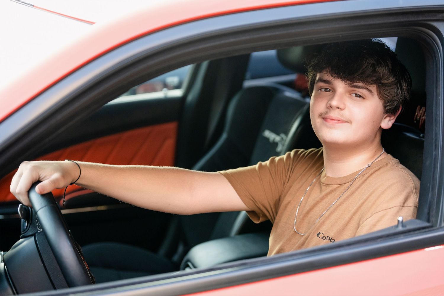 Someone wearing a tan t-shirt sits behind the wheel of an orange sports car.