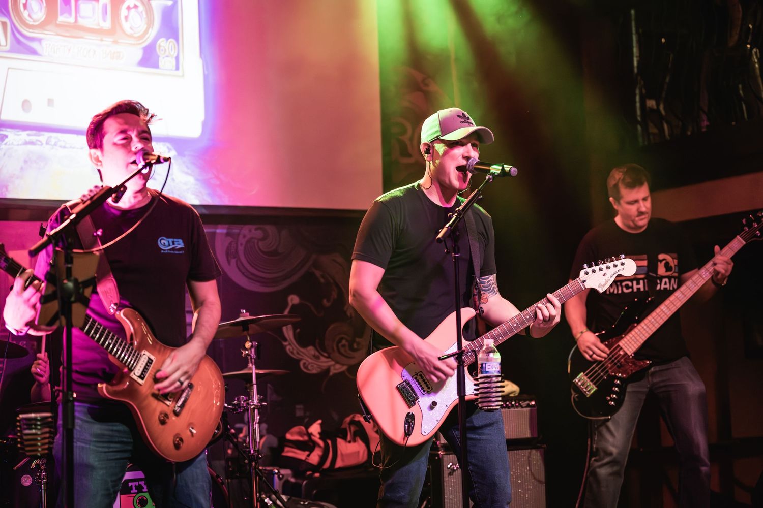 Rock band performing on stage with three guitarists under mixed colored lighting.