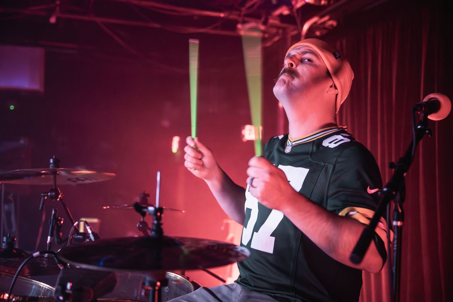 Energetic drummer performing with drumsticks raised under red stage lighting.