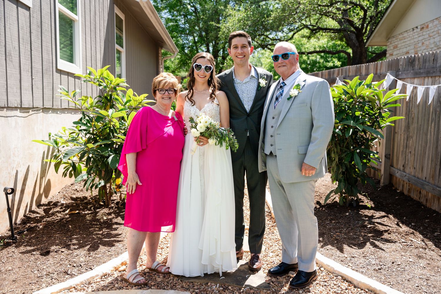 A wedding party poses for a formal portrait outside a rustic building on a sunny day.