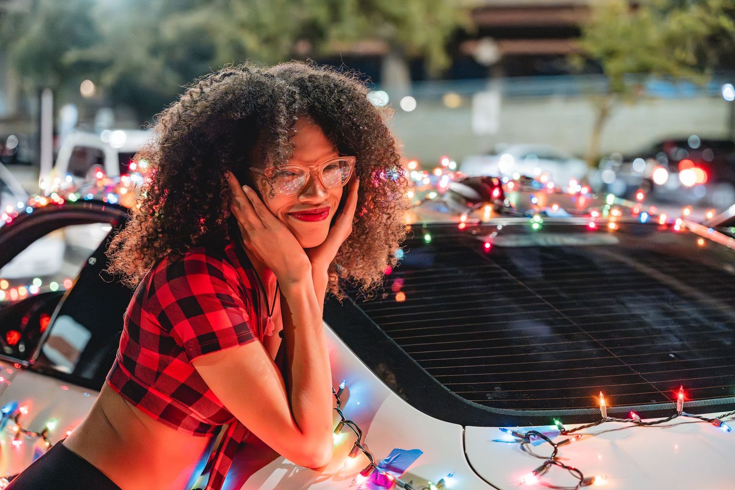A person in plaid crop top poses playfully with holiday lights on car hood.