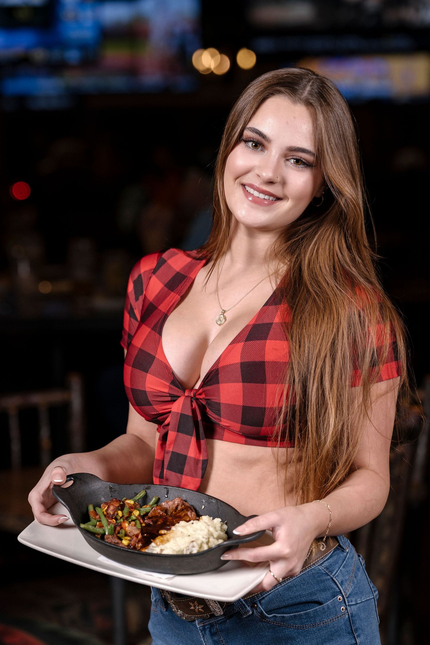 Server in buffalo plaid crop top presents a plate of food at restaurant.