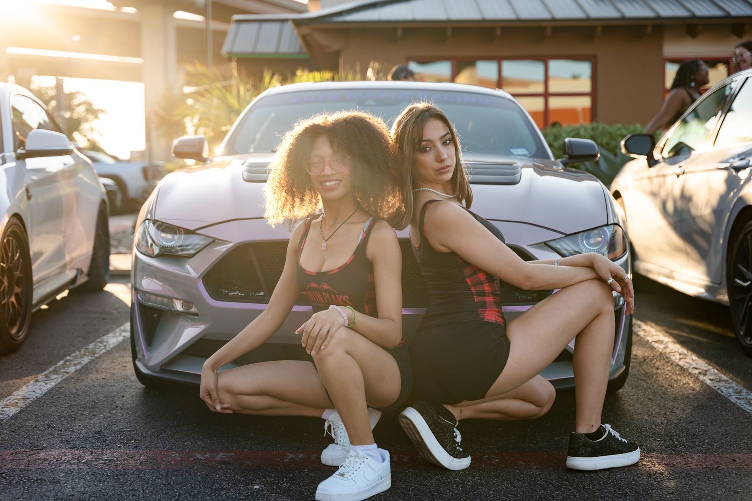 Two friends in casual attire posing in front of a parked car during golden hour.