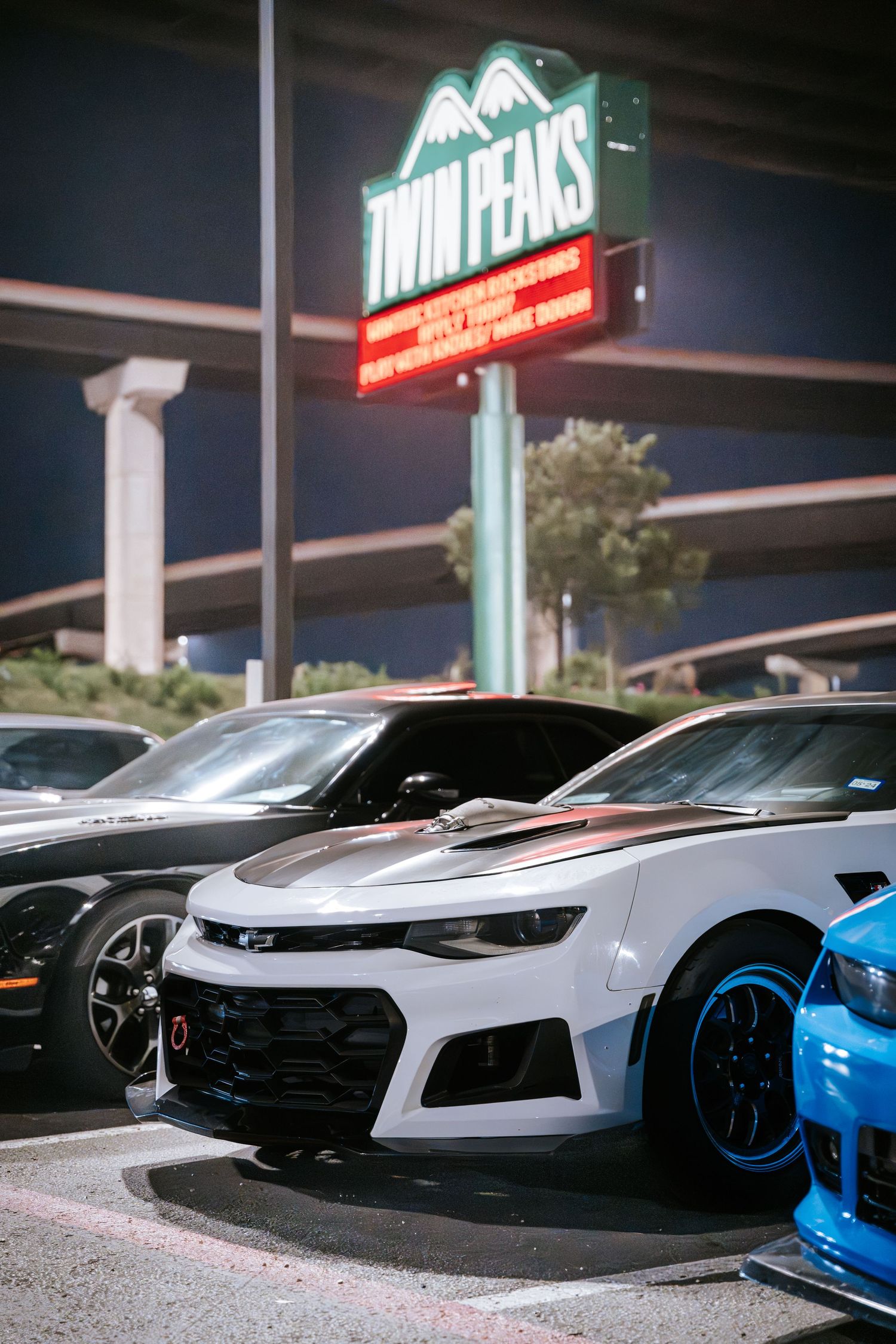 White and blue performance cars parked outside Twin Peaks restaurant at night.