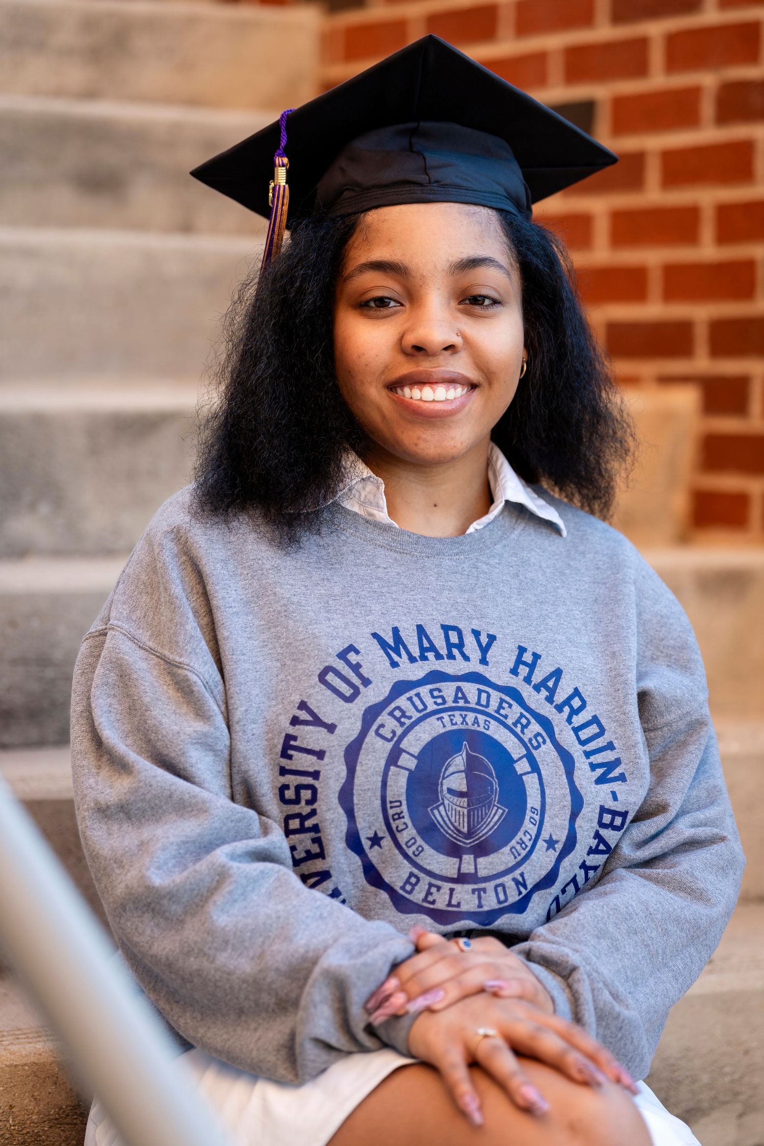 A graduation photo against brick backdrop showing university sweatshirt and traditional black cap and tassel.