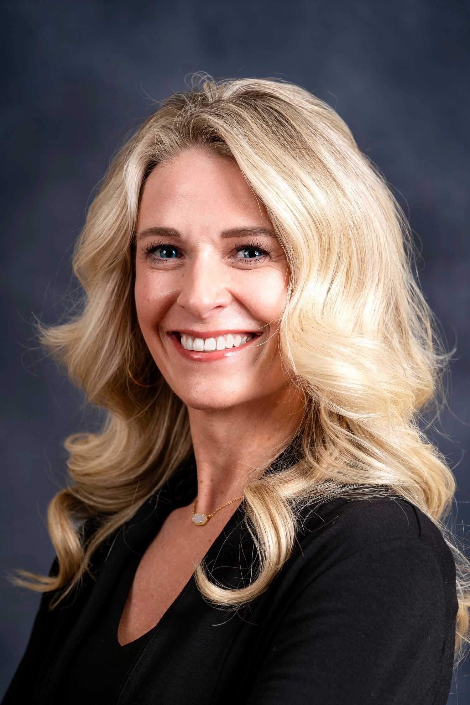 Professional headshot of a smiling person with blonde wavy hair wearing a black blazer against a dark background.