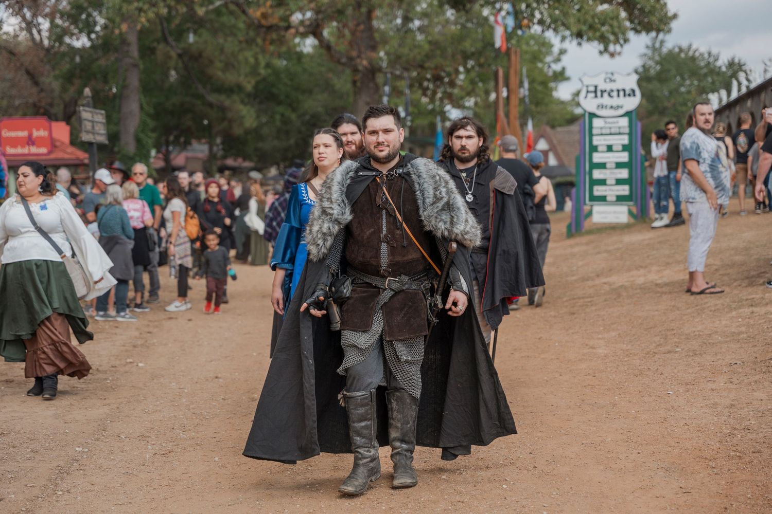 Festival attendees in medieval cloaks and costumes walking along dirt path.