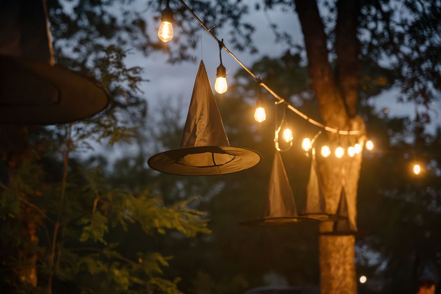 Witch hat decorations hanging from string lights at an outdoor evening party.