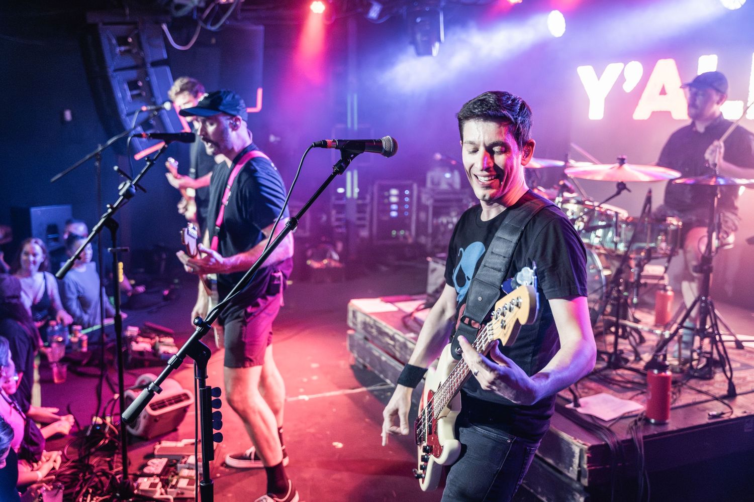 Musicians perform energetically on stage with guitars and drums under colorful purple and red lighting during a live concert.