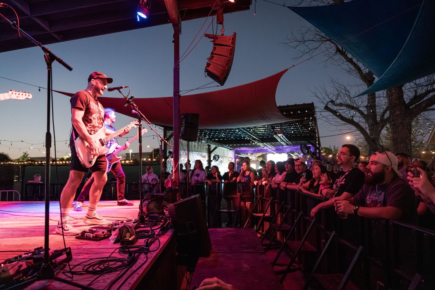 Guitarist performs on outdoor stage at dusk before a large crowd under colorful lights and red tent canopy.
