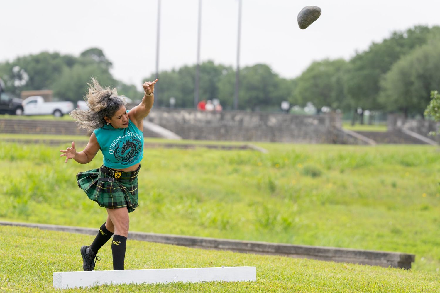 Young girl in tartan skirt and green top throws a stone at a Highland Games competition on a grassy field.