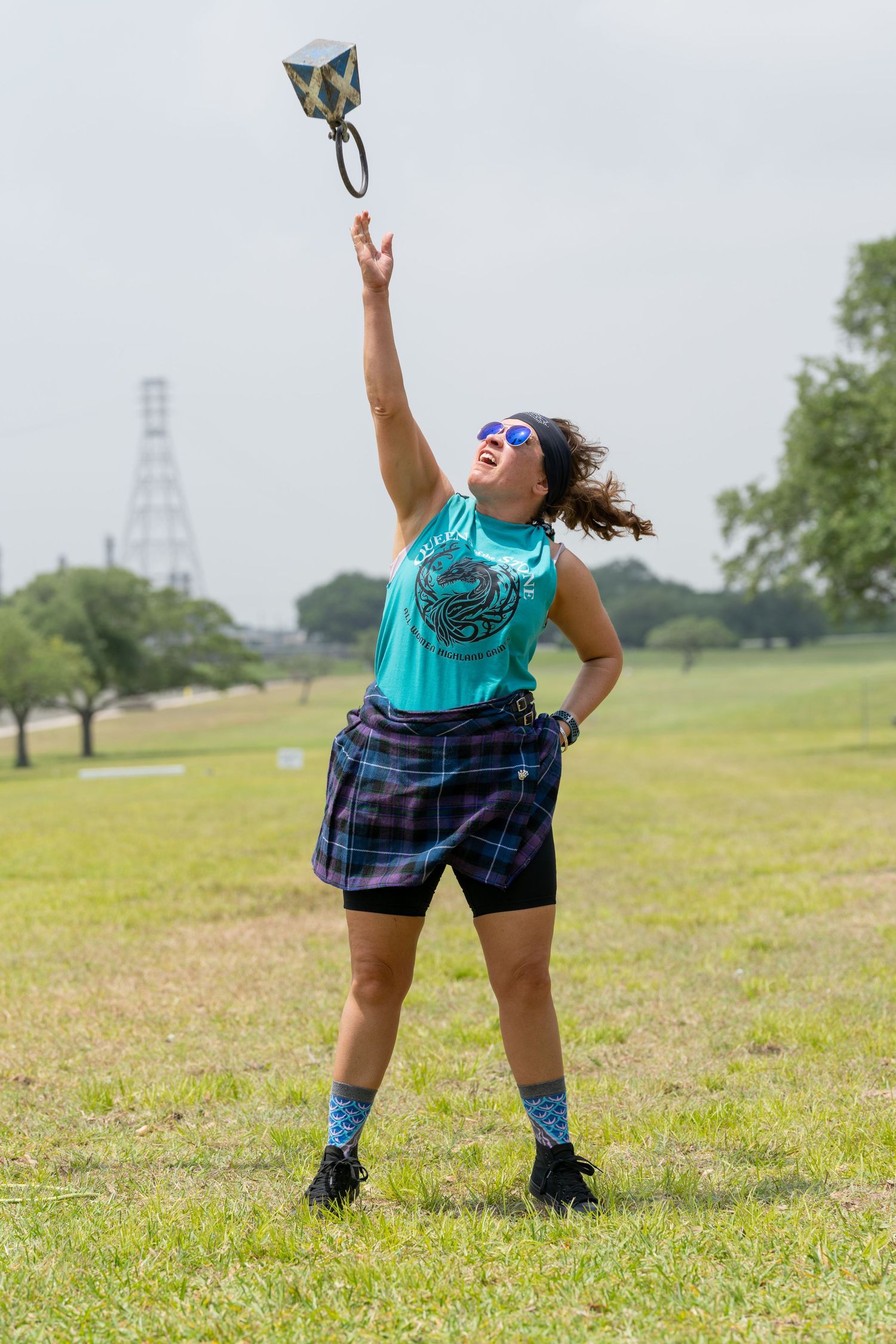 Woman in teal tank top and plaid skirt tossing a weight into the air at an outdoor Highland Games competition.