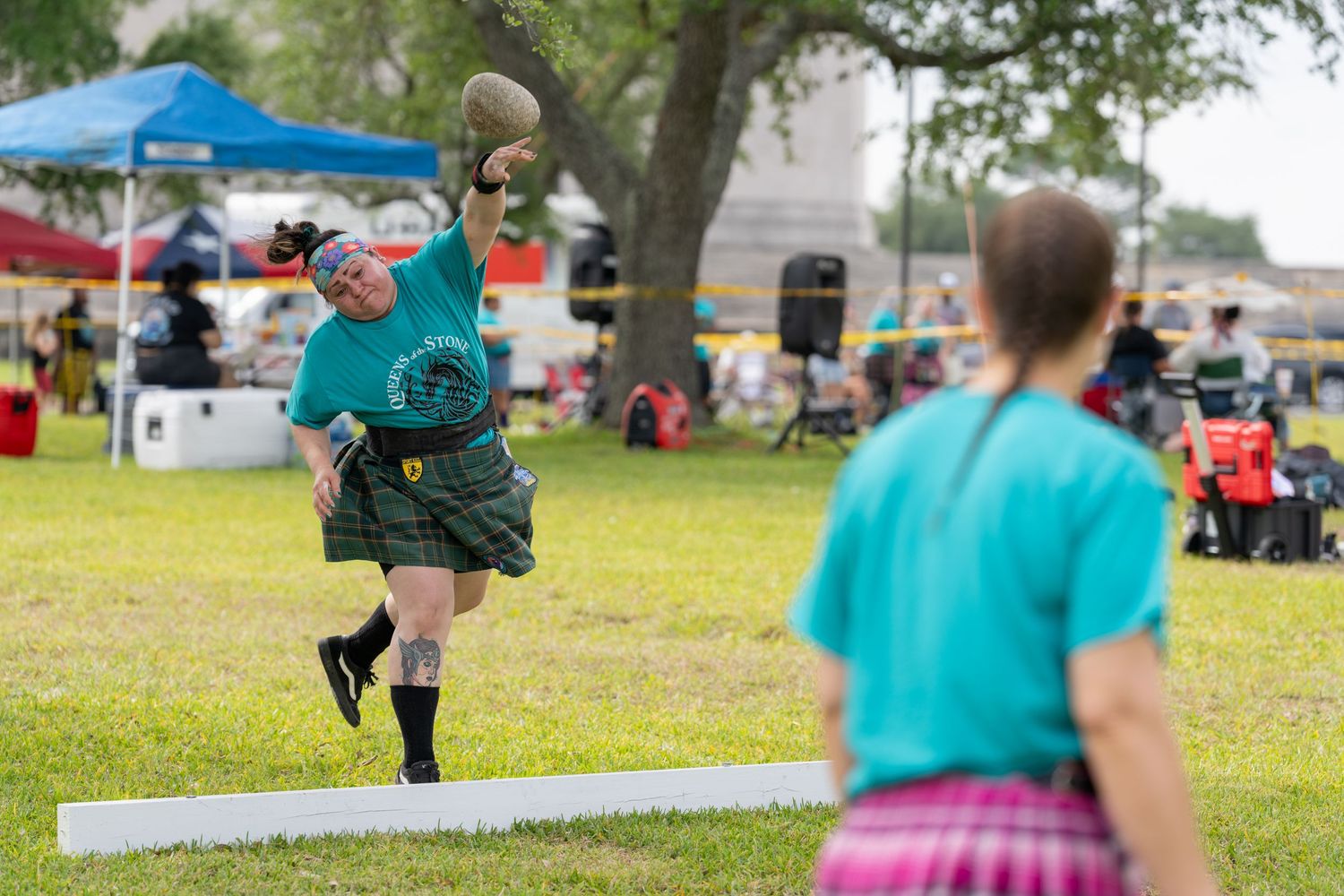 Two athletes in kilts compete in Highland Games heavy events, with one throwing a stone at an outdoor festival.