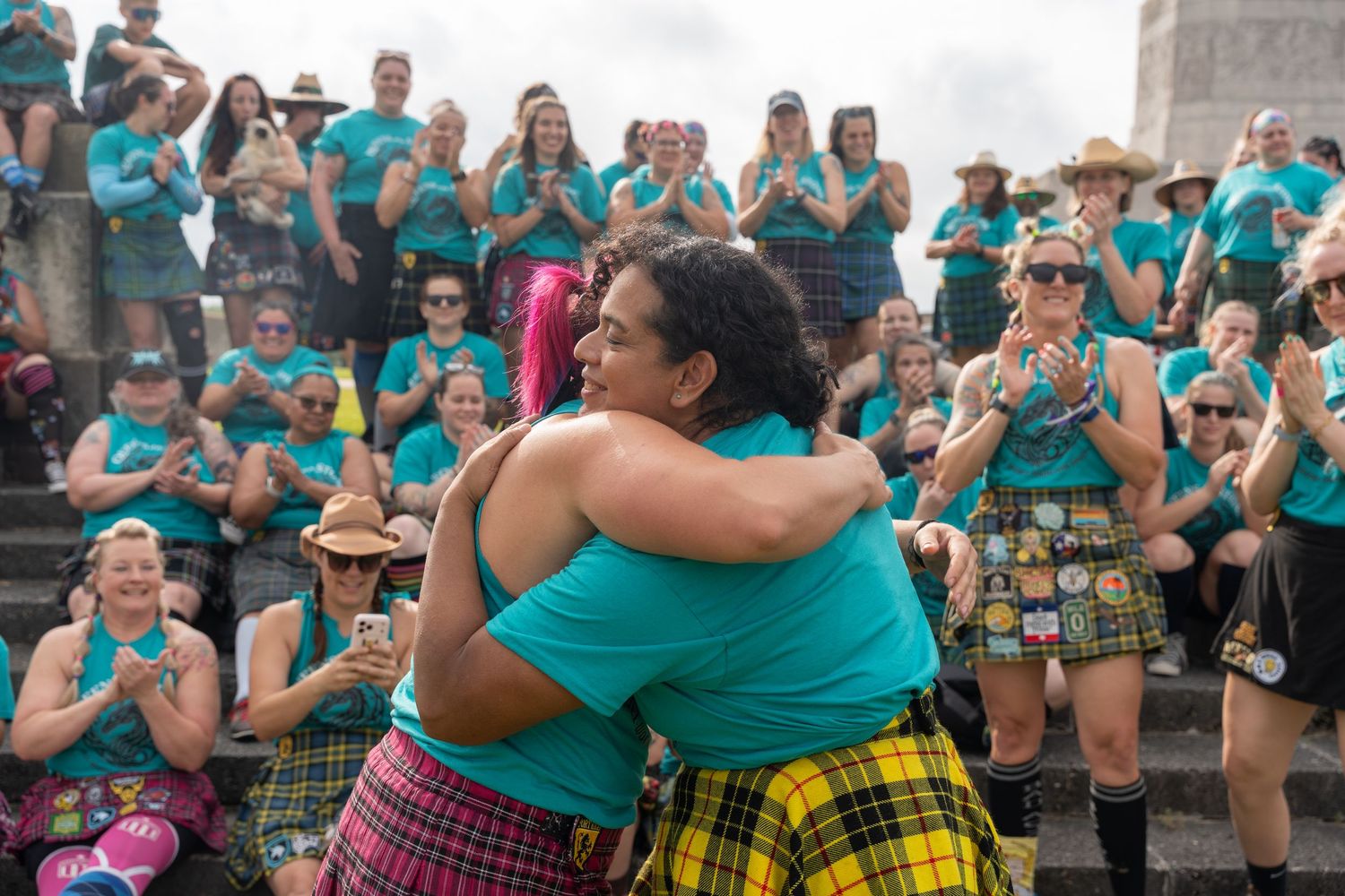 Two women in teal shirts and plaid kilts share a warm hug while a crowd cheers at an outdoor gathering.