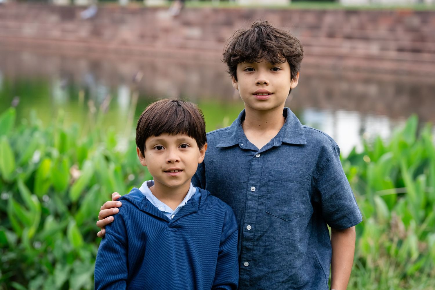 Two young boys in blue shirts pose together outdoors near green plants and a brick wall.