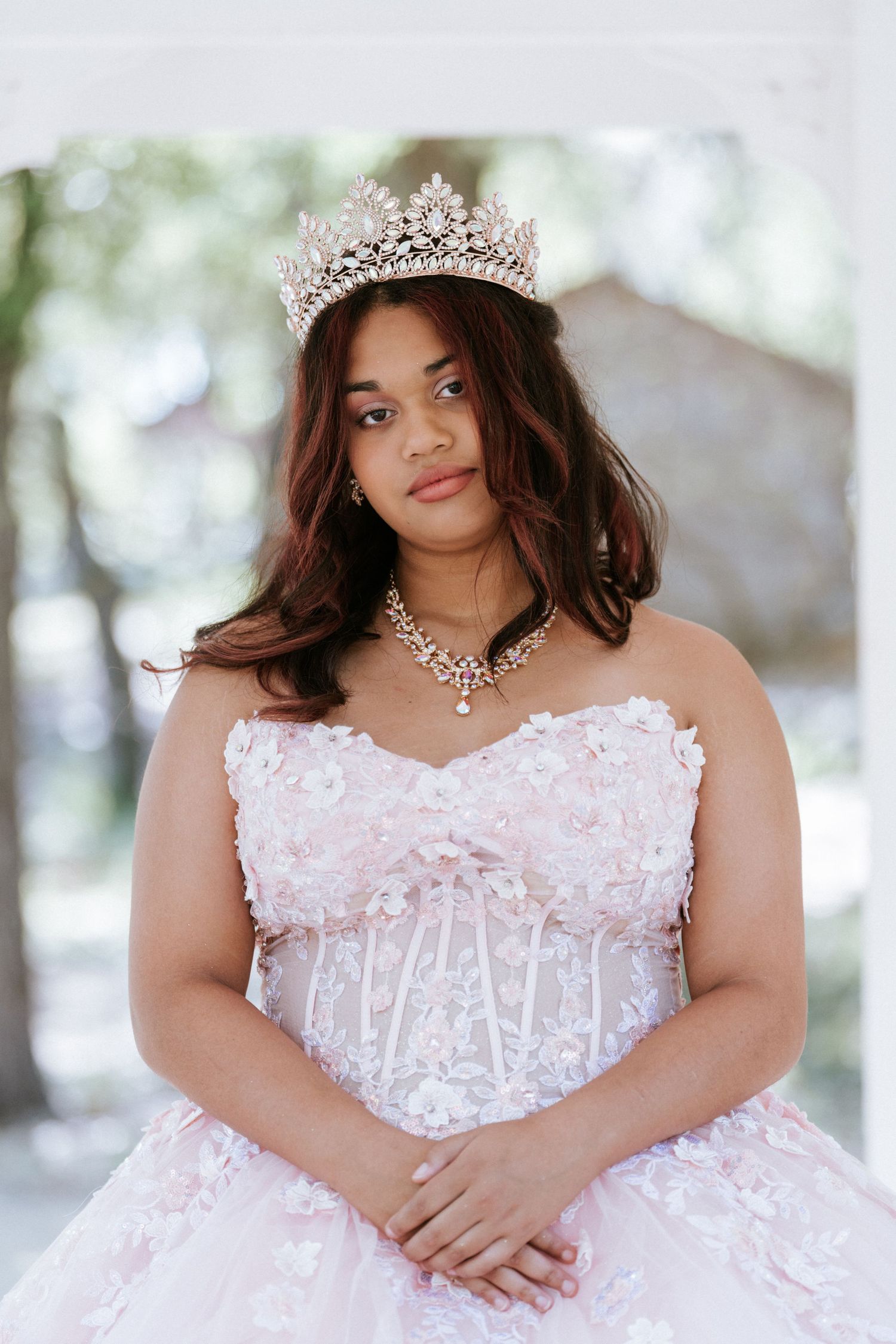 Young woman in pink floral ball gown and crystal crown poses elegantly for quinceañera portrait outdoors.