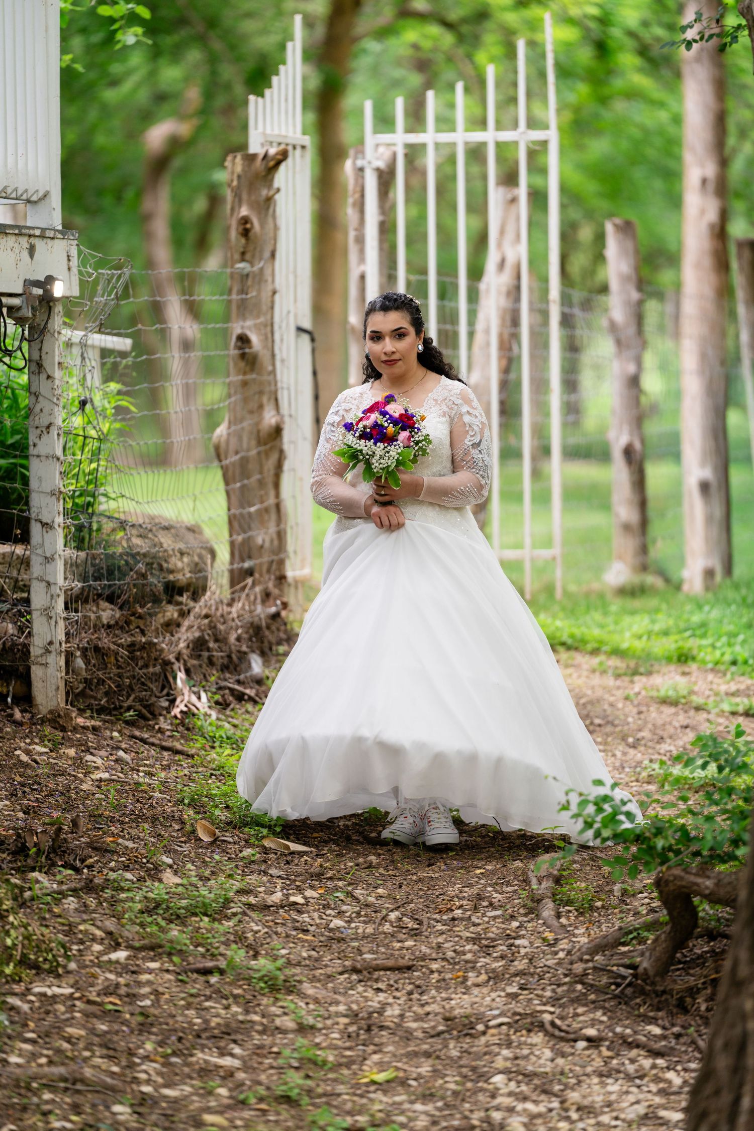 Bride in white lace ball gown holding colorful bouquet stands on garden path near white gate surrounded by trees.
