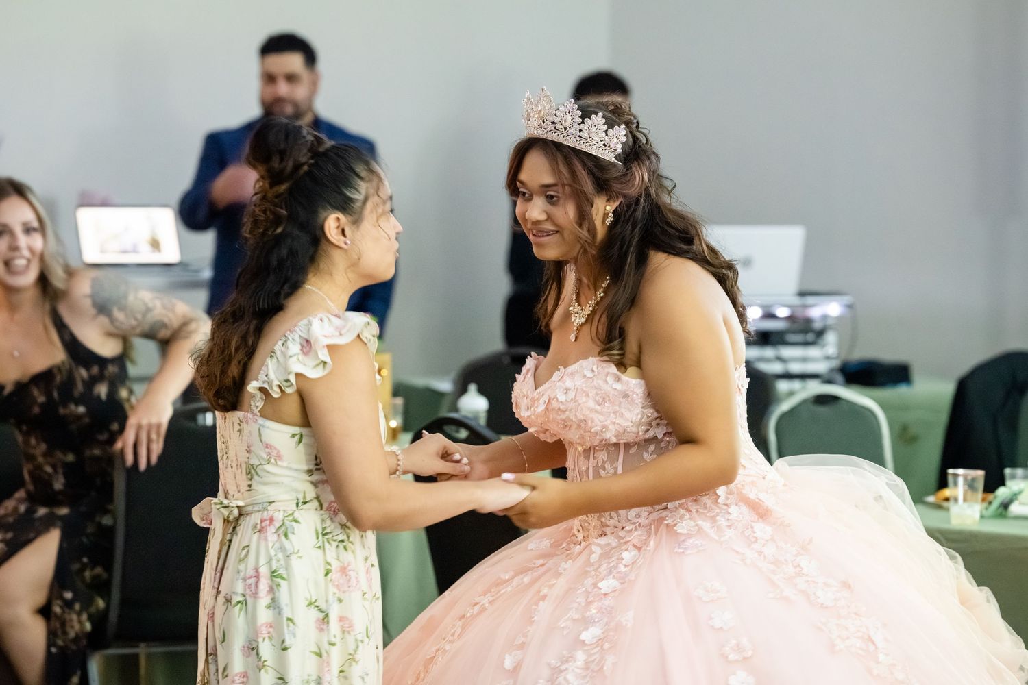 A girl in a pink quinceañera gown and tiara shares a joyful moment with a younger girl at a celebration.