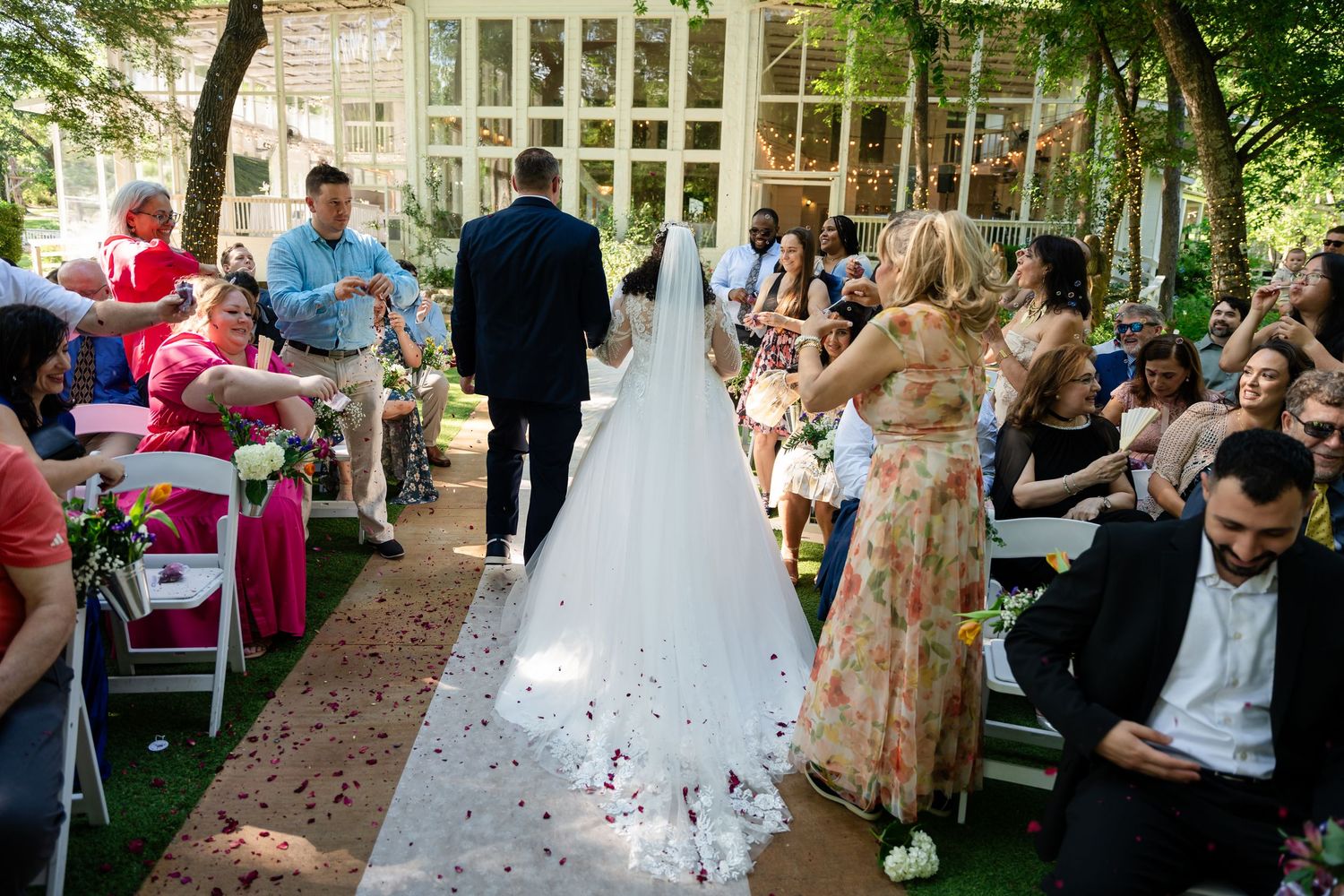 Bride in white gown walks down outdoor aisle with long veil, surrounded by seated wedding guests in a garden ceremony.