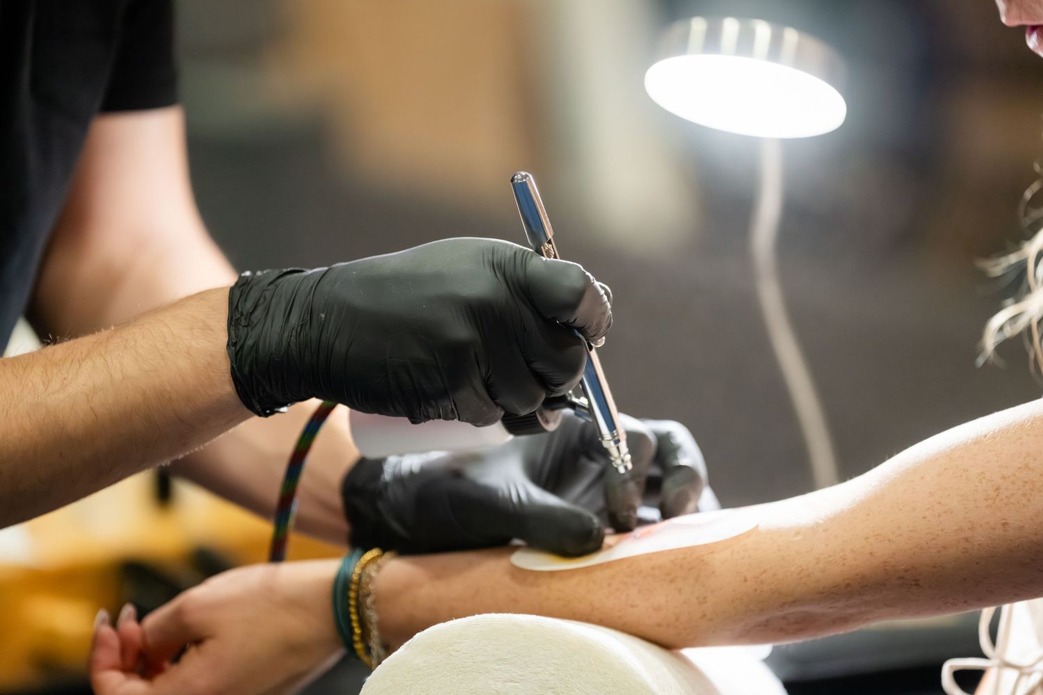Tattoo artist wearing black gloves carefully applies a tattoo needle to a client's arm under a bright lamp.
