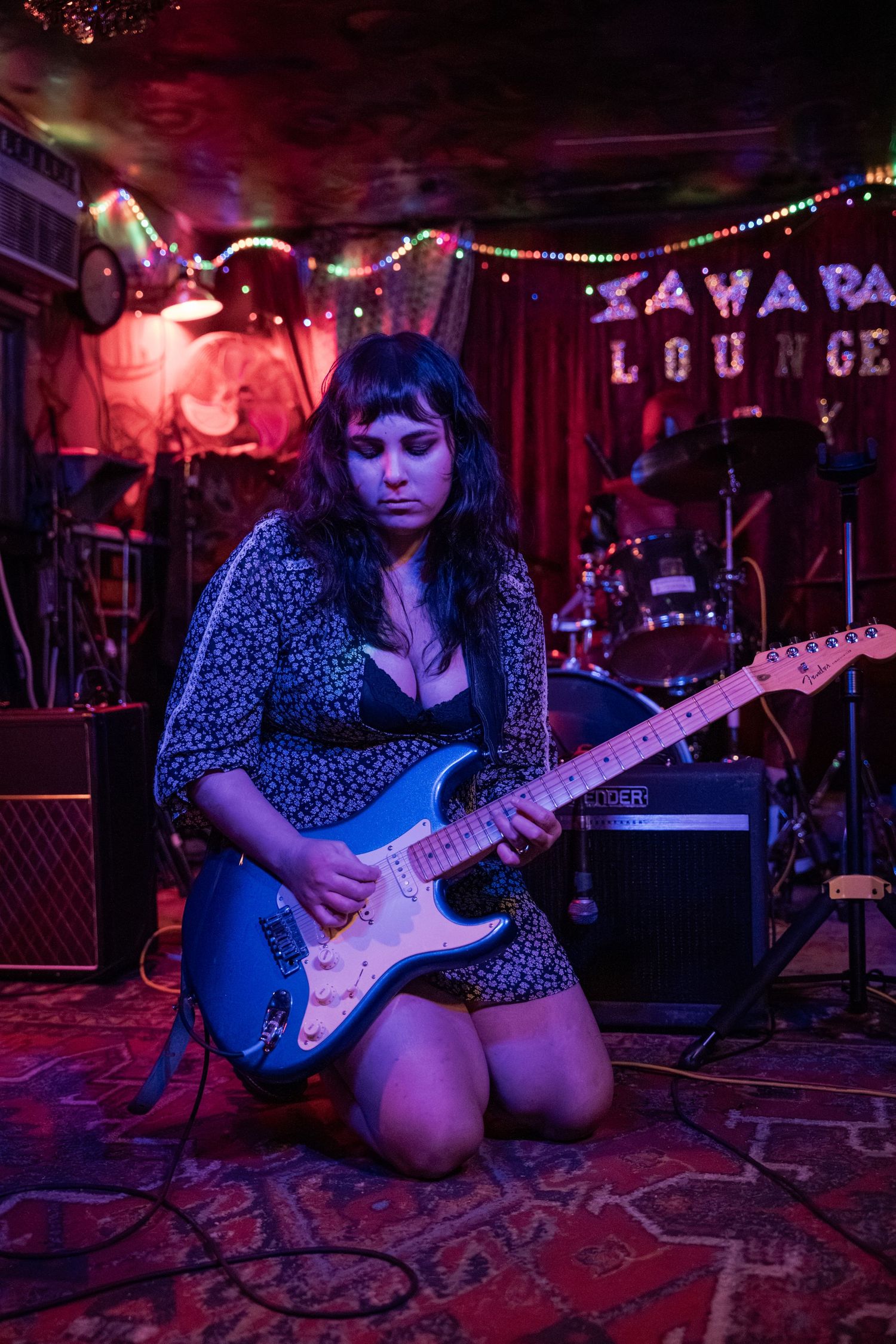 A musician kneels on stage playing an electric guitar at a dimly lit lounge venue with neon lights and string decorations.