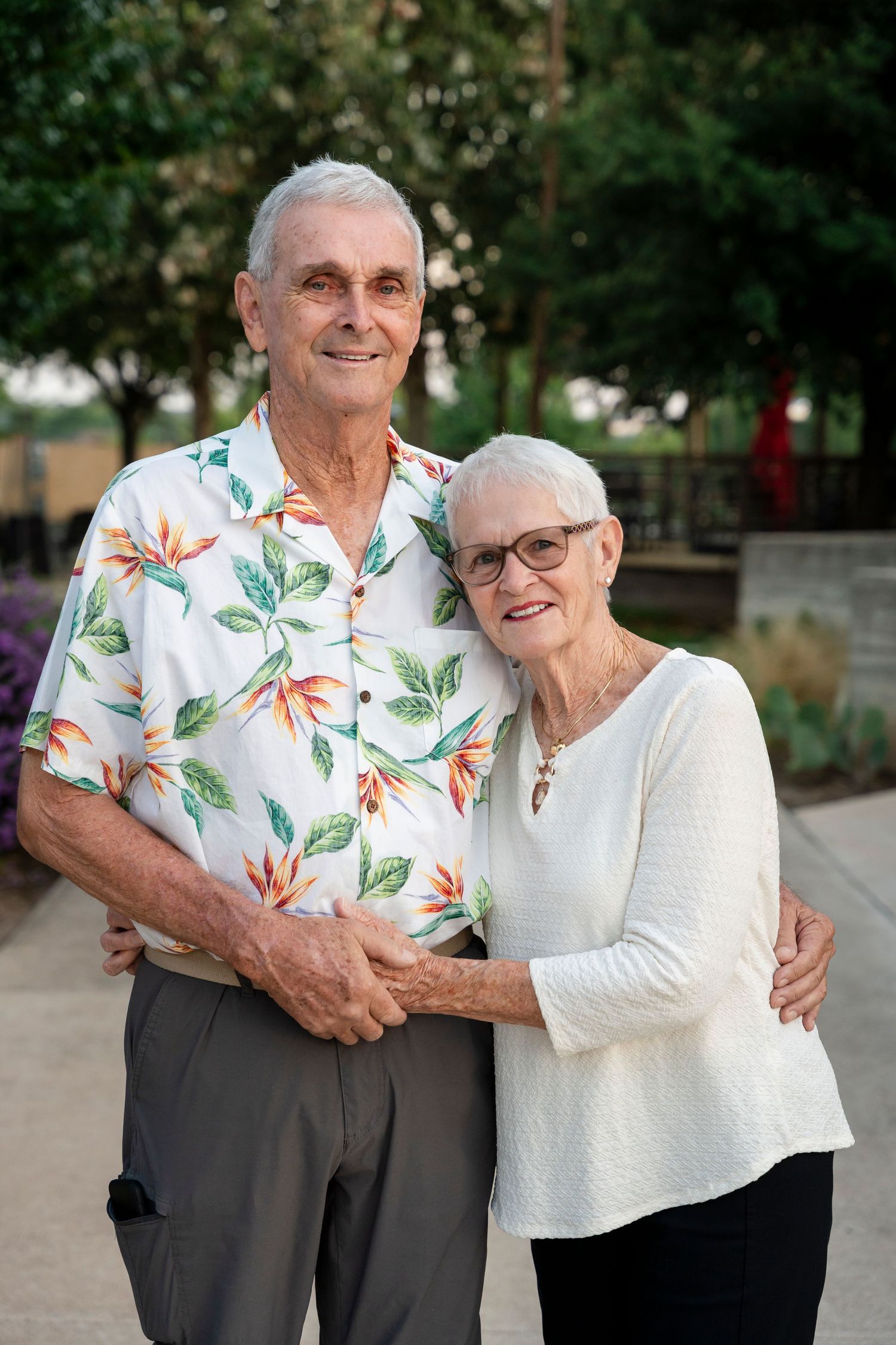 An elderly couple embraces outdoors while wearing coordinated outfits with floral patterns and white clothing.