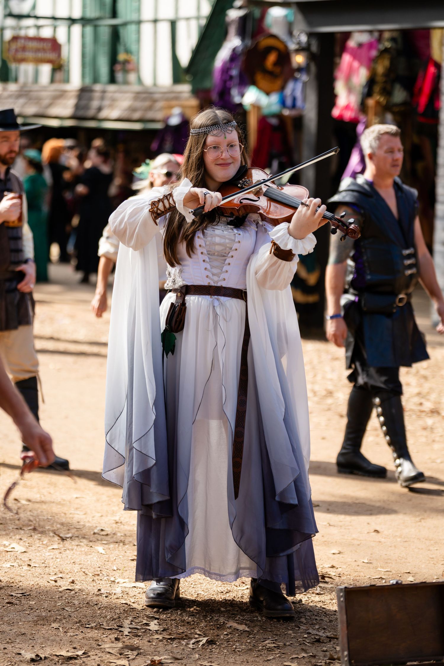 Three medieval costumed performers in flowing blue and white robes walk together at a renaissance festival.