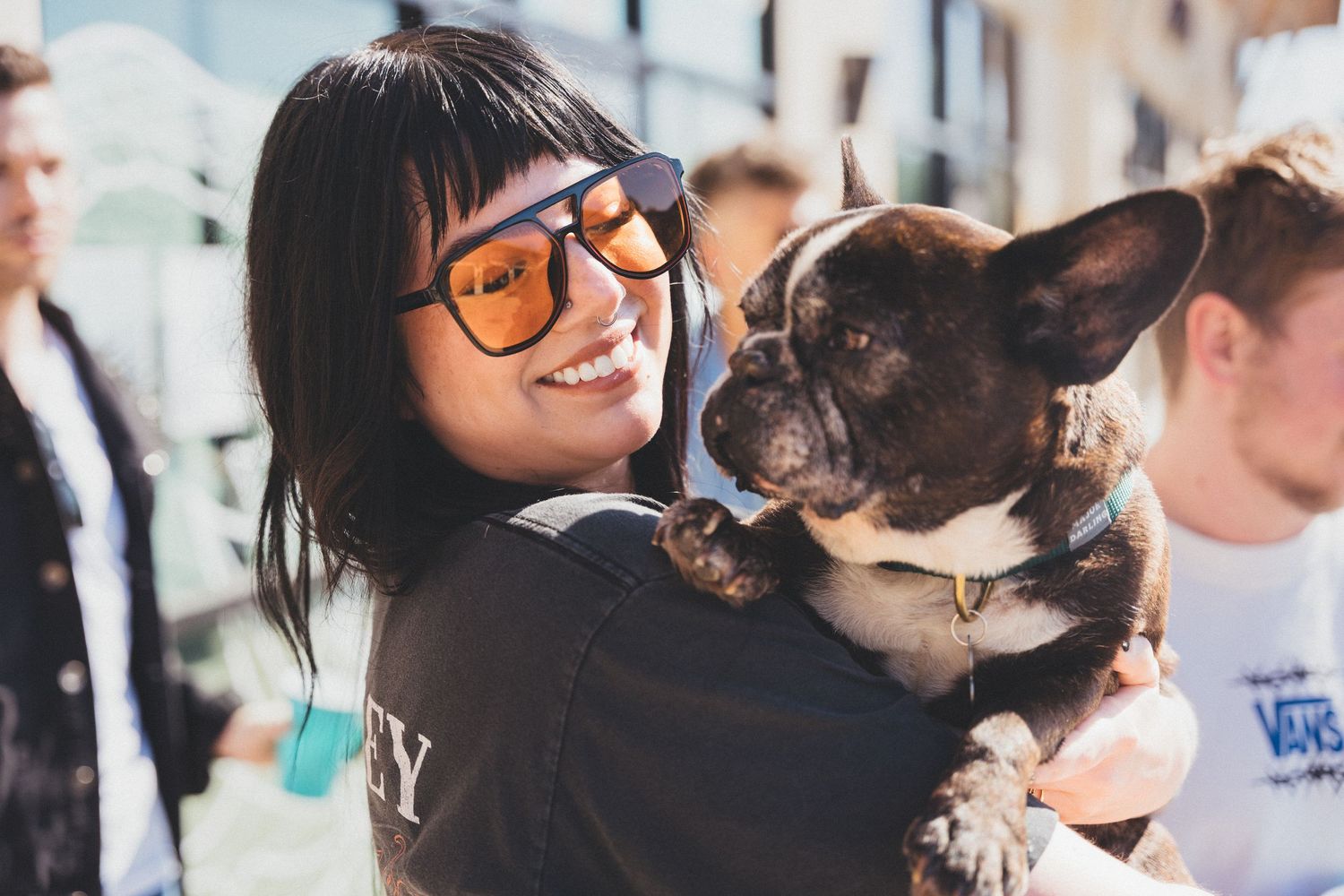Person wearing glasses holds and smiles with a French Bulldog outdoors on a sunny day.