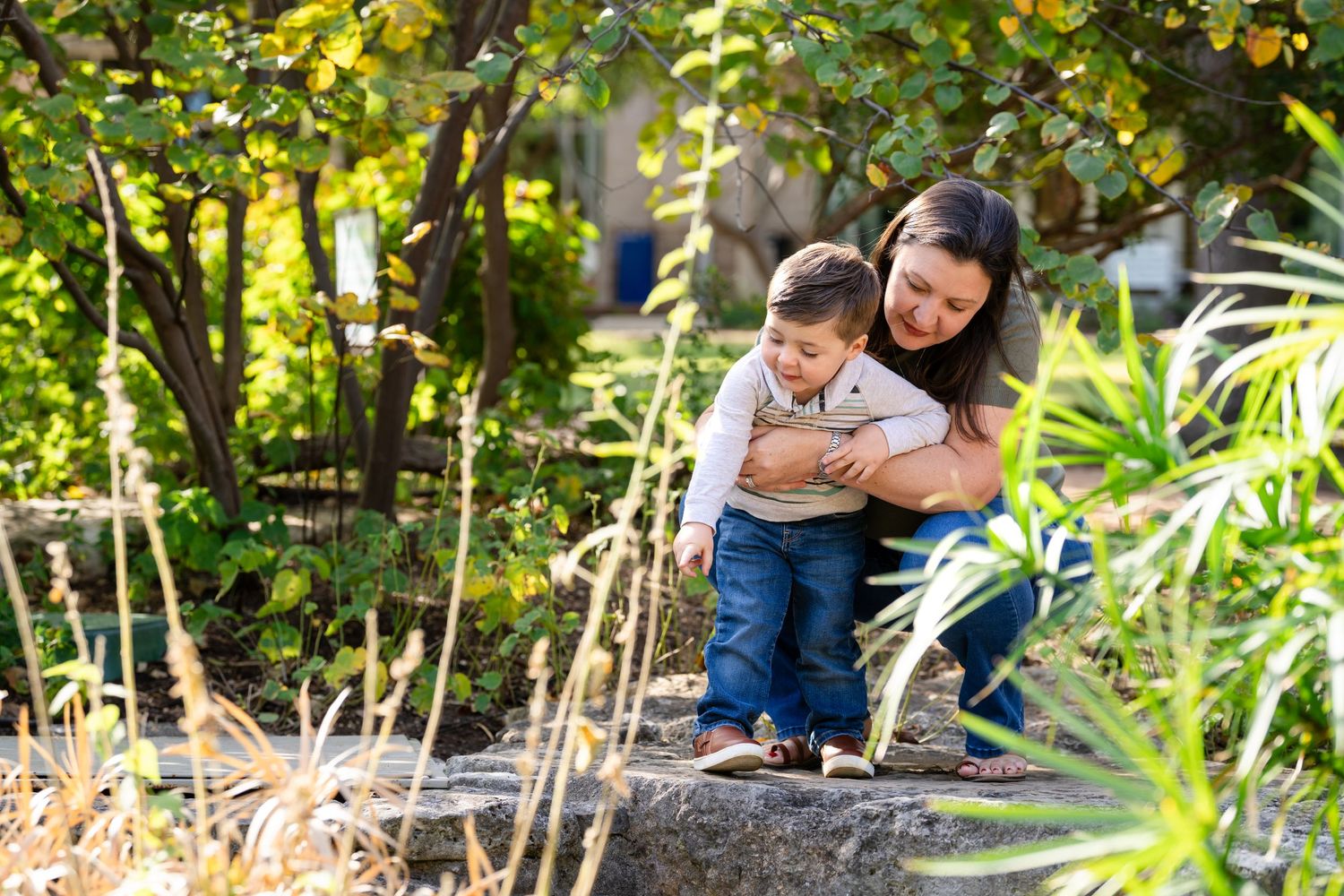 Two people share a tender embrace while sitting together in a lush garden setting with dappled sunlight.
