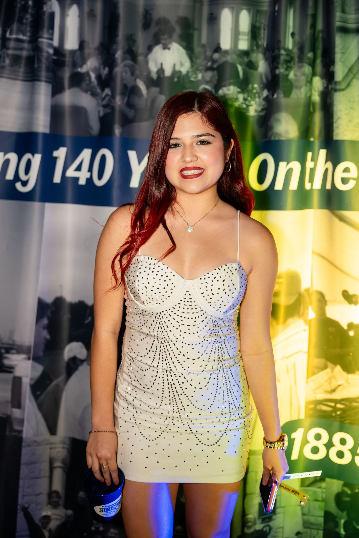 A woman in a white beaded mini dress poses at an event with colorful lighting and backdrop.