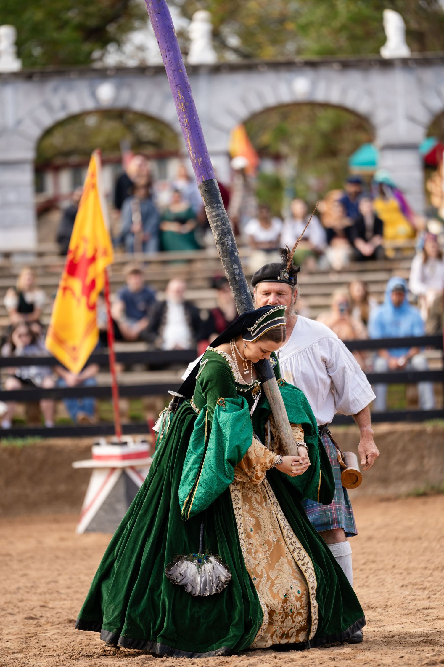 Performers in medieval costumes engage in sword fighting at a Renaissance fair in emerald green and gold dresses.