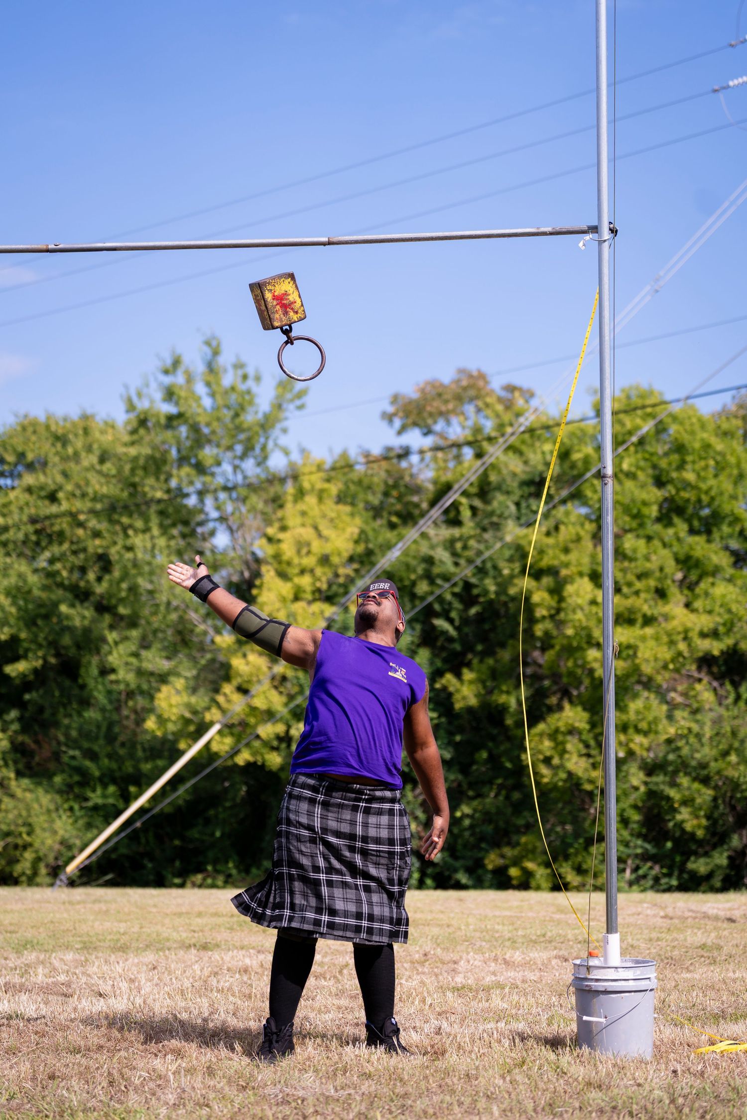 Athlete in purple shirt and plaid kilt throws weight during Highland Games competition on sunny field.