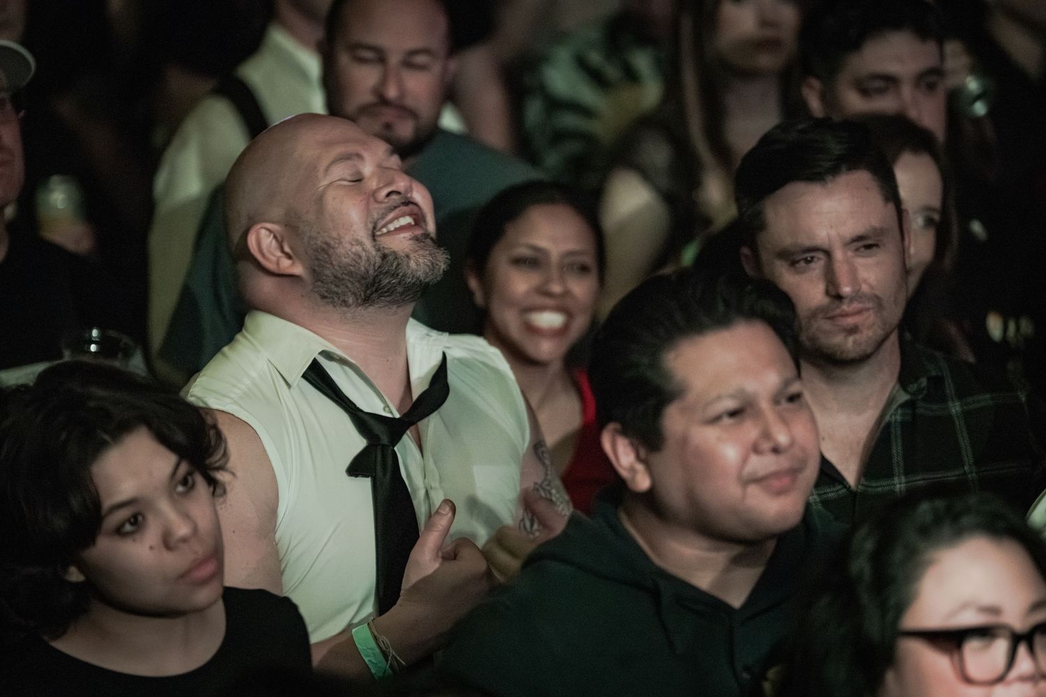 Crowd of diverse people at a concert, man in green shirt with black tie laughing joyfully.