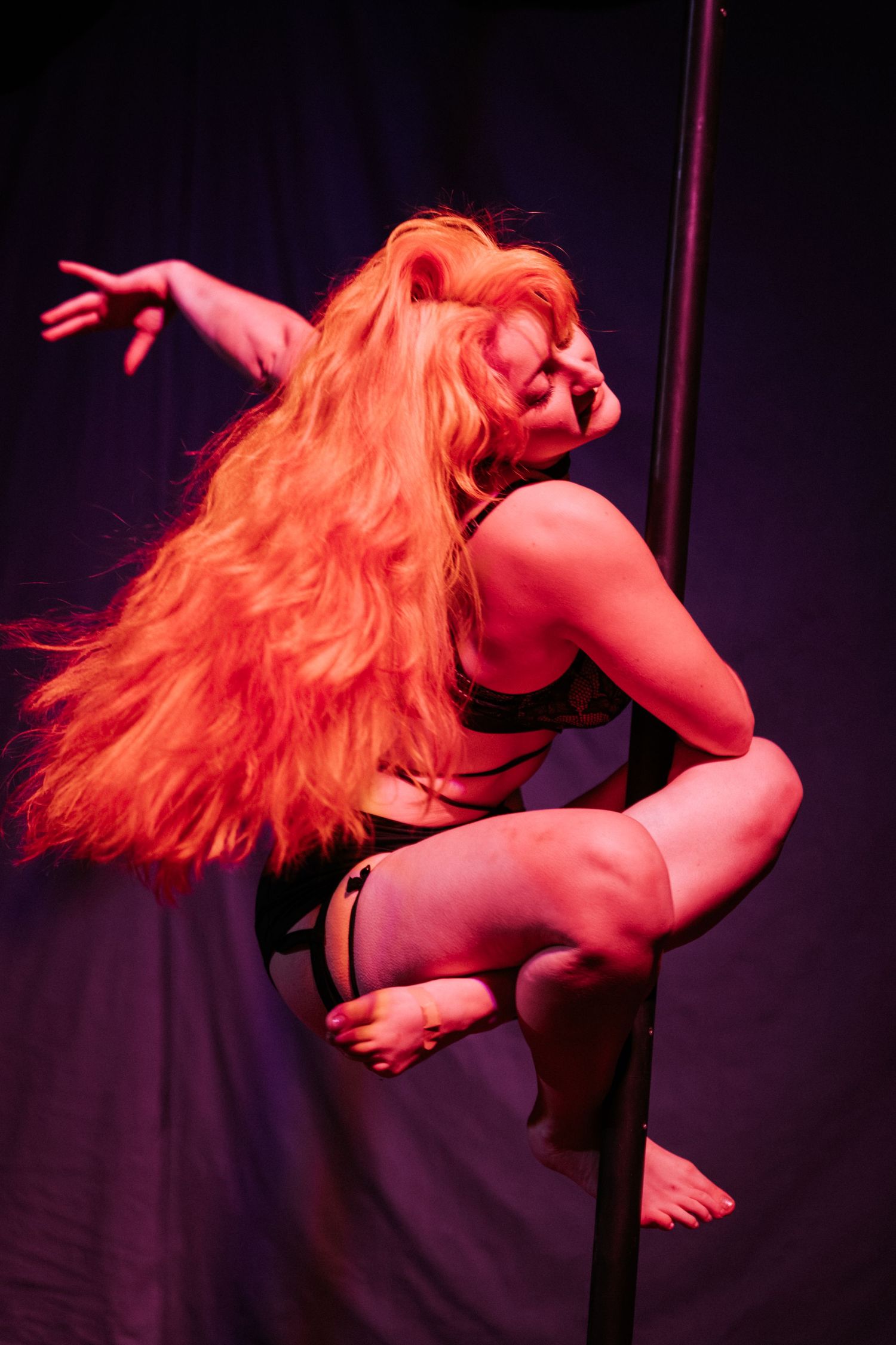 Red-haired woman performing pole dance under dramatic red stage lighting in black lingerie.