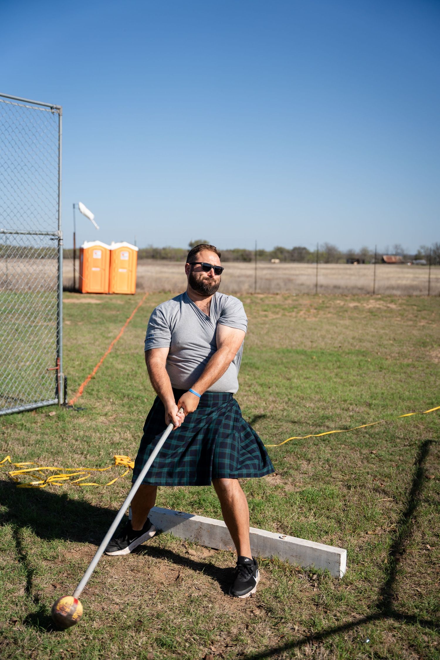 Man in tartan kilt and sunglasses competing in Highland Games, holding a caber on a grassy field.