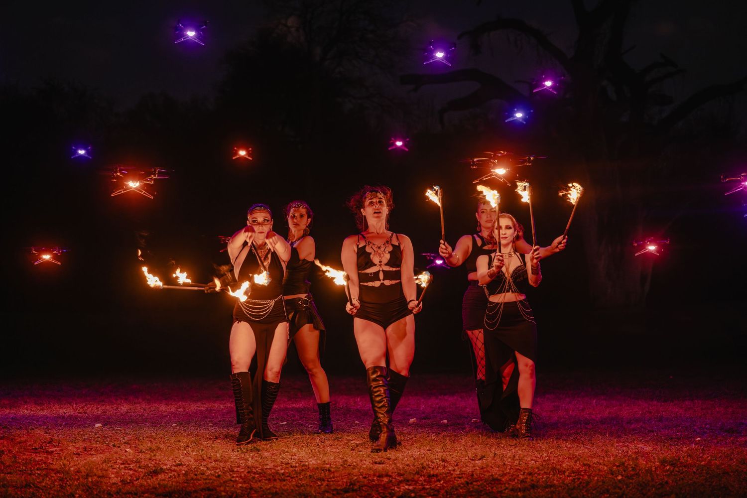 Five fire performers in black costumes pose dramatically at night holding flaming props with purple lights in background.