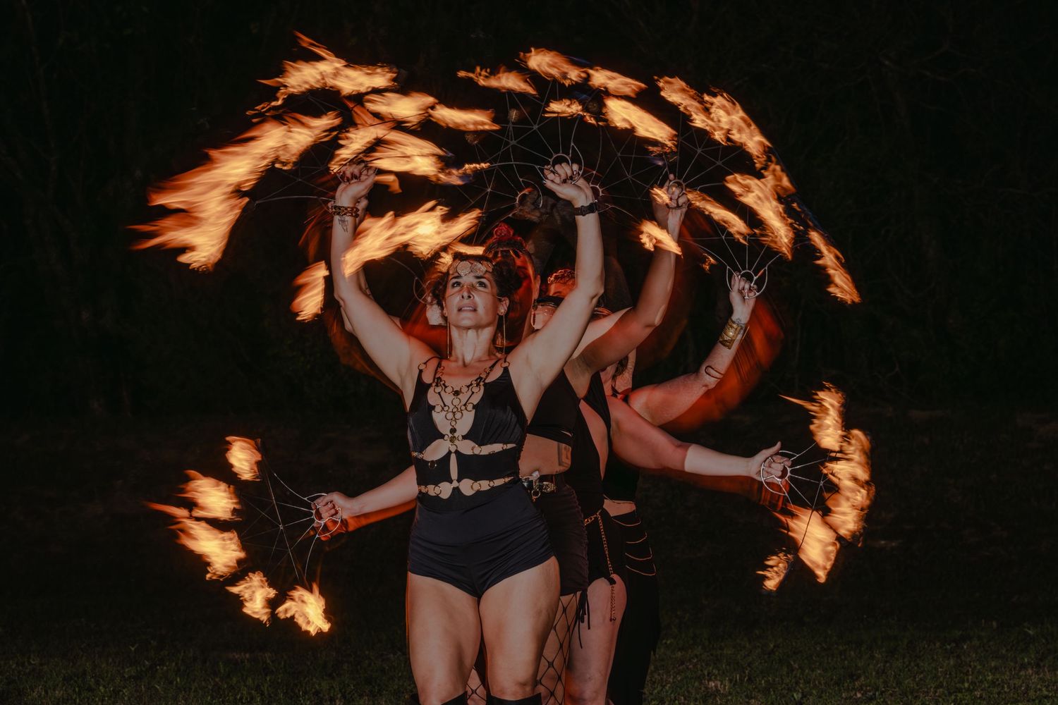 Fire dancer performs stunning flaming poi routine at night outdoor event, wearing black costume with multiple fire props.