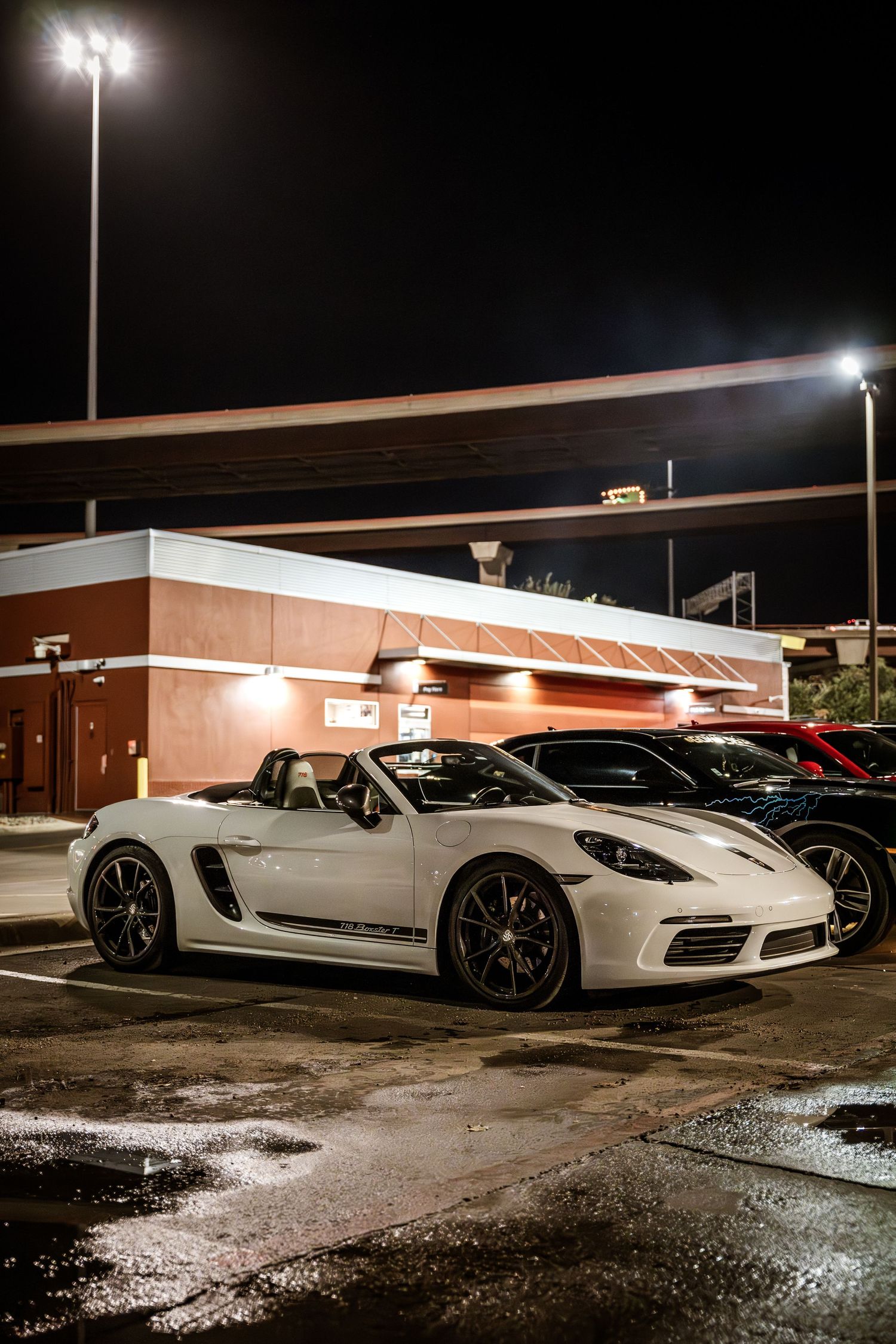 White Porsche Boxster with aftermarket wheels parked under bright parking lot lights at night.