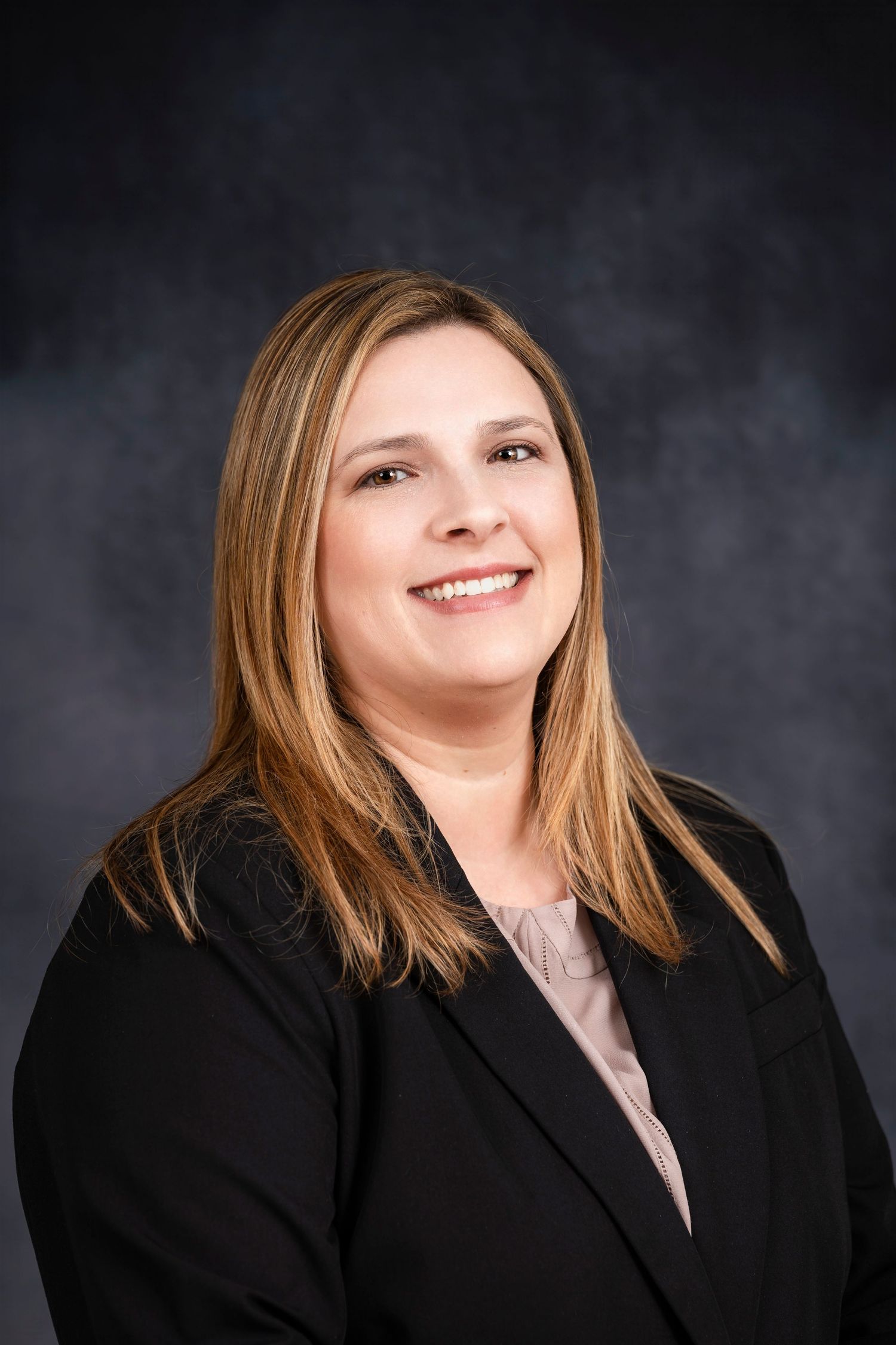 Professional business portrait of a woman wearing a black blazer against dark backdrop.