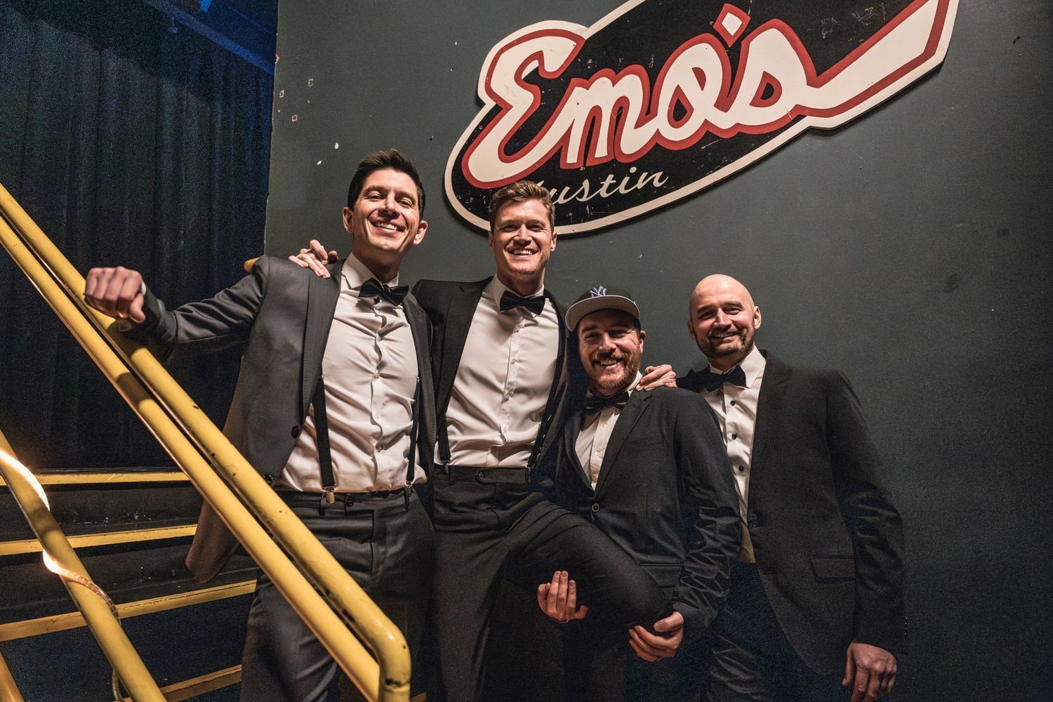 Band members in suspenders and bowties pose on a staircase at Emo's venue.