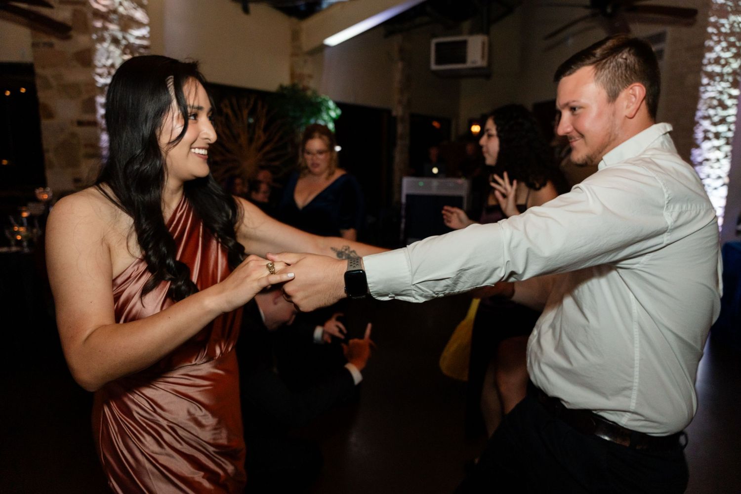 Wedding guests dance joyfully together in a brown dress and white shirt on a dimly lit dance floor.