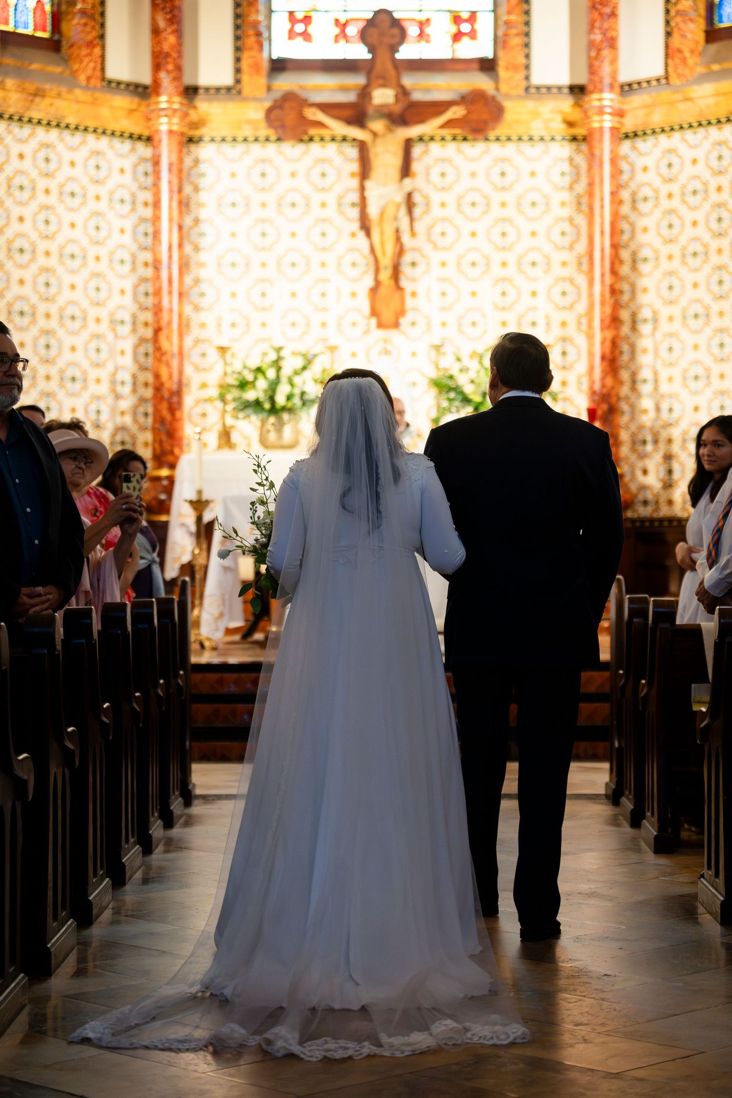 Bride and groom stand at altar during Catholic church wedding ceremony with crucifix and decorative wall patterns visible.